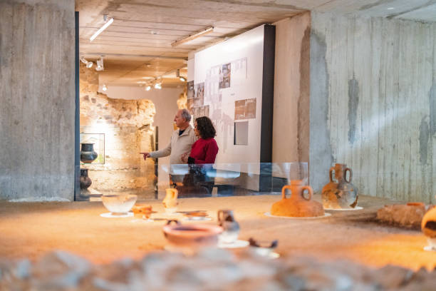 A senior couple visiting an interesting history exhibition in a museum. They are looking at old vases and artefacts displayed in the displays cases. The senior man is pointing at an old vase while talking to his wife.
