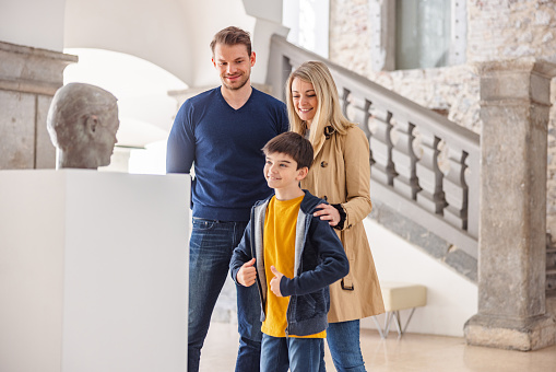A young caucasian family visiting a history museum on the weekend. The parents are attractive and young- They have light hair. They are happy to be taking their son to a museum. The brunette young boy looks happy and enthusiastic. The family is standing close to a statue of a man's head located on a pedestal in the centre of the room. The museum is beautiful and bright.