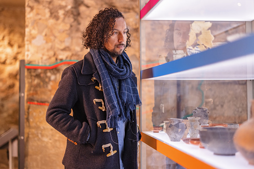 A focused adult black male looking at a display of of artefacts belonging to older cultures and civilisations. He is standing next to a glass display cabinet and looking into it. He is visting a history museum during the weekend. The man has long curly hair and a beard. He is wearing  a blue scarf.
