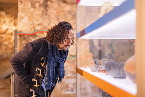 An adult black male leaning forward in order to see the display of artefact better. The artefacts are protected glass. In the display there are various old pottery vessels. The man looks interested in what he is looking at at the museum.