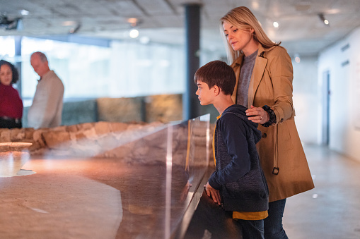 A caucasian mother taking her son to a history exhibition in a museum. The mother and her young son are looking through glass in order to see the artefacts displayed. The mother and son are serious and focused. The mother has her hand on the boy's shoulder. The boy looks interested.