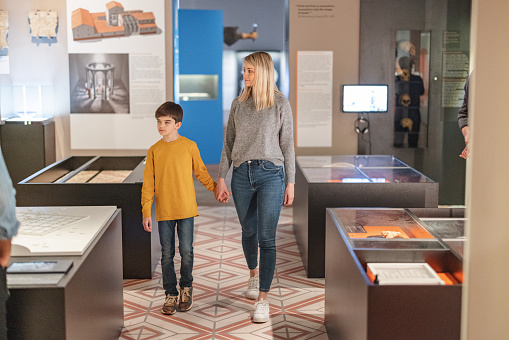 A blonde caucasian single mother visiting a history exhibition in a museum with her young brunette son on a weekend. She and her son are looking around while holding hands. They are walking past tje different display cases with artefacts.