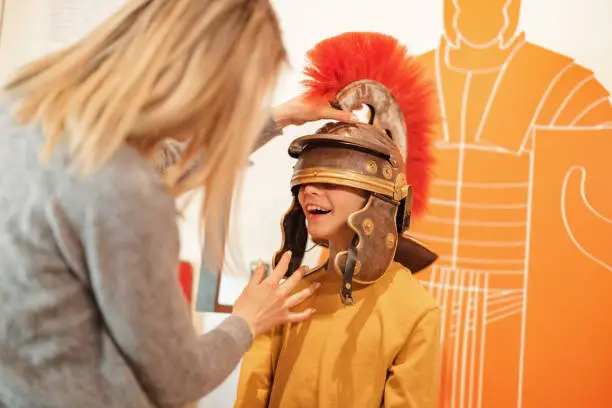 A blonde caucasian mother putting on a Roman galea on her happy young son's head during their visit of the Roman Empire exhibition. The mother and her son are visiting a museum on the weekend. The son is smiling as he is standing with a large helmet on his head. He is unable to see.