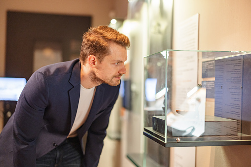 An adult blonde caucasian male museum visitor looking at a small statue displayed in a small glass display case. The male is attractive and is wearing classy clothes. He is visiting a history museum and looks fascinated by the artefacts displayed.