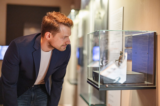 An attractive adult caucasian male leaning forward and looking at a display case of a small statue while visiting a museum. He is dressed in a classy jacket. The man looks serious and focused.