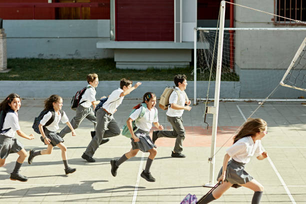 Full length side view of 9, 10, and 11 year old school children in uniforms running with backpacks through sunny campus quad.