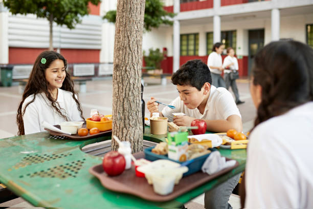 School children enjoying snack time outdoors Waist-up view of 10 and 11 year old classmates smiling and laughing as they sit at table under tree and eat fruit and yogurt. food elementary student healthy eating schoolboy stock pictures, royalty-free photos & images