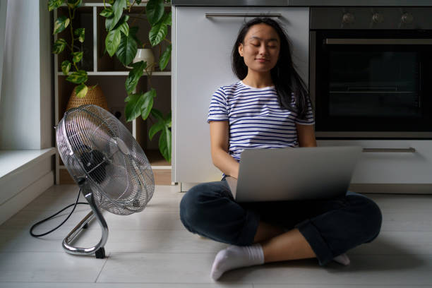 istock Contented Asian woman turn on floor ventilator fan while work with laptop joyful self save from heat 1464720211