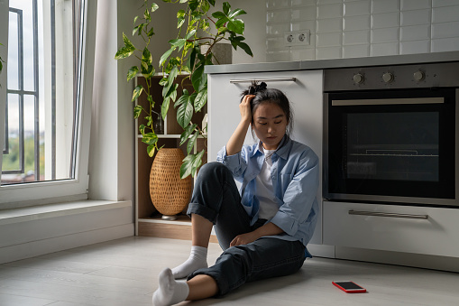 Upset depressed Asian girl suffering from breakup, heartbroken frustrated woman sits on floor in kitchen looking at smartphone waiting for call or message from ex boyfriend. Codependent relationships