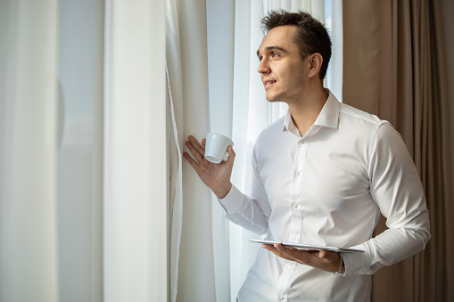 A man dressed in a white shirt stands by the window in a hotel room with a cup of coffee and a digital tablet, portrait of a young businessman, confident, ready for a meeting with business partners