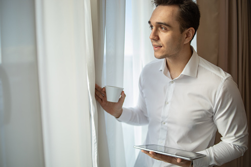 A man dressed in a white shirt stands by the window in a hotel room with a cup of coffee and a digital tablet, portrait of a young businessman, confident, ready for a meeting with business partners