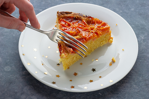 An anonymous woman's hand cutting into a slice of Blood Orange Almond cake with a fork. The piece of cake is on a white plate with gold stars on a blue gray table.