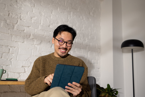 Young hip Chinese man sitting at his home office while enjoying his work using tablet