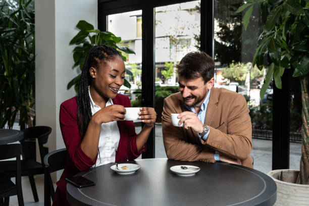 Young man and African American woman sitting in cafeteria drinking coffee flirting talking laughing and seducing outside working hours and colleague in restaurant away from busy street stock photo