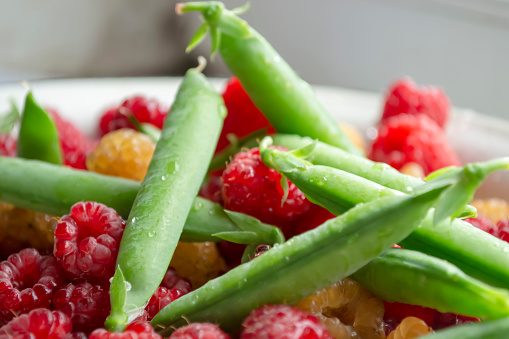 Fresh, summer berries - green peas in pods and red raspberries. Green peas are in the same bowl with red raspberries. Healthy legumes are in the summer.