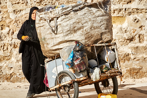 Sousse, Tunisia, February 8, 2023: Elderly woman in black abaya and hijab pushes a handcart with a sack in which she collects plastic waste for recycling