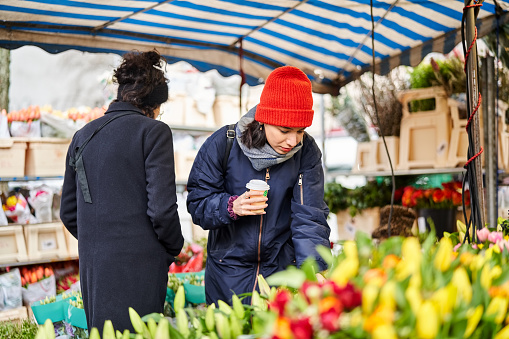 Young woman in warm clothes holding a coffee looking at flower in local market stall. Woman shopping flowers at flea market in the city during autumn.