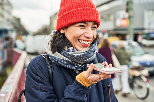 Close-up of cheerful young woman talking on speaker phone outdoors in city. Female in warm clothing using cell phone in the city on autumn day.