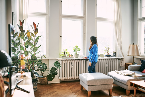 Full length of mid adult woman standing by radiator and looking outside the window at home. Mid adult female standing in living room looking out of window.