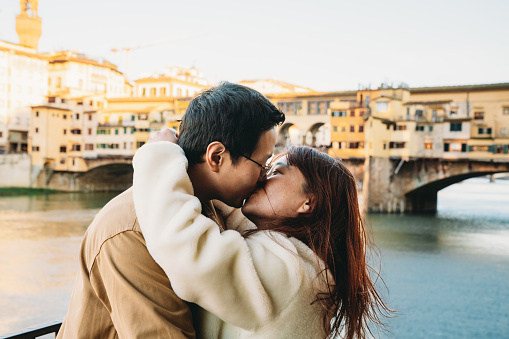 An asian couple is kissing in Florence with Ponte Vecchio in the background. They are exploring the city together during their honeymoon.