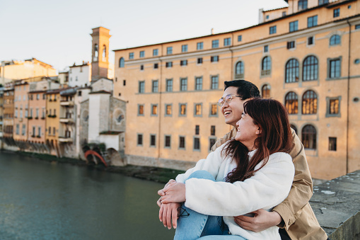 An asian couple is sitting on Santa Trinita bridge in Florence, enjoying the sunset together. They are traveling in Europe for their honeymoon.