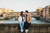 An asian couple is sitting on Santa Trinita bridge with Ponte Vecchio in the background - Florence, Italy