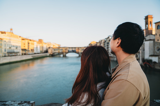 An asian couple is embracing on Santa Trinita bridge with Ponte Vecchio in the background - Florence, Italy. They are enjoying their honeymoon in Europe together.