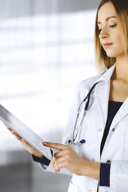 istock Young woman-doctor is holding a tablet computer in her hands, while standing in a clinic. Portrait of friendly female physician with a stethoscope at work. Perfect medical service in a hospital. Medicine concept 1464741326