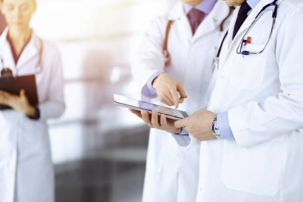 istock Group of unknown doctors use a computer tablet to check up some medical names records, while standing in a sunny hospital office. Physicians ready to examine and help patients. Medical help, insurance in health care 1464741374
