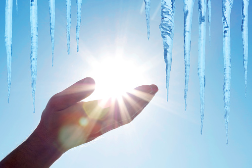 Close-up of melting icicles and a hand as if holding the sun with beautiful sunbeams. Blue sky background. Space for copy.