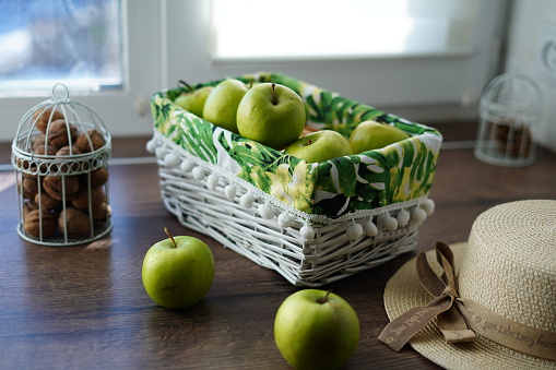 A white wicker basket with green juicy apples on a wooden table near the window. Next to the basket lies a beige hat and a walnut basket. Sunny day. Beautiful background