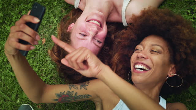 Portrait of Two Multiethnic Women Lying Down on a Park Lawn Using a Smartphone and Laughing. Diverse Young Female Bestfriends Enjoying the Sunny Warm Weather Outside, Scrolling Through Videos