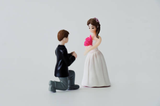 Wedding and marriage theme - Miniature wedding dolls isolated on white background, focus on bride. Groom kneels down, putting ring onto bride's finger; selective focus