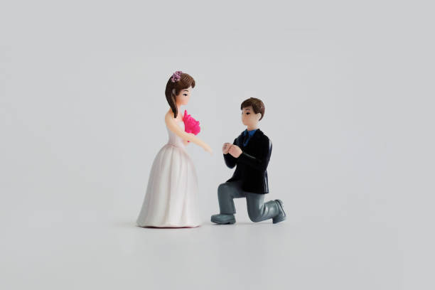 結婚式のテーマ – 白い背景にミニチュアの結婚式の人形。新郎はひざまずき、花嫁の指に指輪をはめます。
