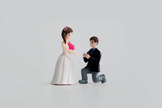 Wedding theme - miniature wedding dolls isolated on white background. Groom kneels down, putting ring onto bride's finger