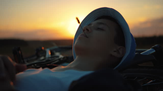 Funny teenager boy relax outdoors, lies near the bike at sunset in the field, cinematic shot, steadicam shot