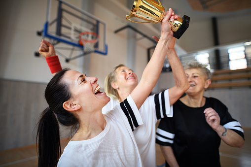 A group of young and old women, basketball team players, in gym with trophy celebrating victory.