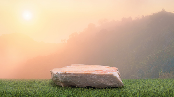 Stone podium table top on fresh green grass with outdoor mountains scene nature landscape at sunrise blur background.Natural beauty cosmetic or healthy product placement presentation pedestal display.