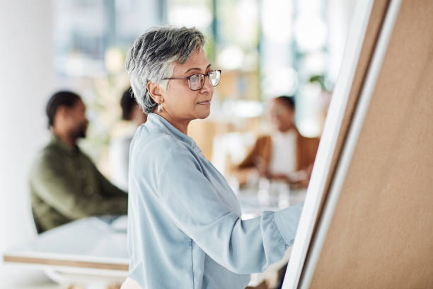 istock Creative senior woman, meeting and writing on Flipchart for team building, coaching or activity at office. Elderly female in presentation using board to write idea strategy or FAQ for company startup 1464764057