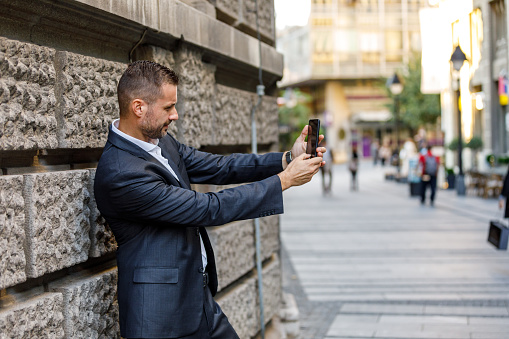A Modern Business Person is Standing in the City Streets and Using his Mobile Phone to Take a Self Portrait Photography.