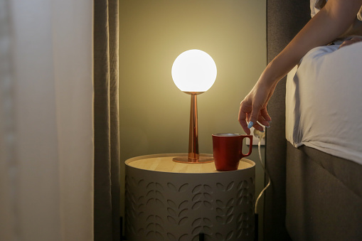 Woman in bed reaching for her first morning coffee from the nightstand. A red mug on the bedside table