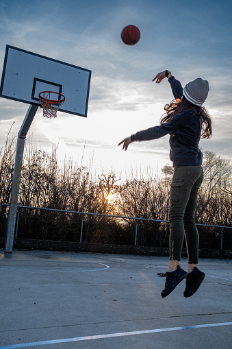 Rear view of a teenage girl, in mid-air, takes a shot on a basketball court, backlit sunset, vertical composition, copy space