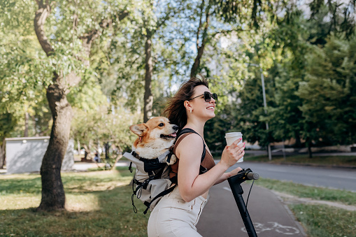 Happy smiling woman traveler is standing with electro scooter and drinking coffee in city parkland with dog Welsh Corgi Pembroke in a special backpack