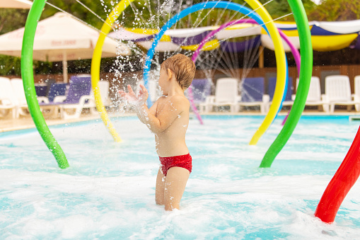 Happy little child stands under the fountain having fun with the water splashing in the pool in amusement park