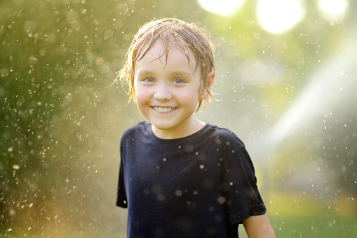 Close-up portrait of funny little boy playing with garden sprinkler in sunny backyard. Elementary school child laughing, jumping and having fun with water. Summer outdoors activity for children.