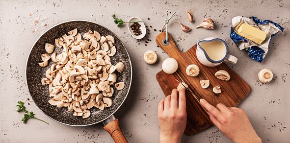 Cooking - preparing creamy mushroom sauce on a pan. Chefâs hands cut recipe ingredients on a wooden board. Kitchen worktop captured from above (top view, flat lay).