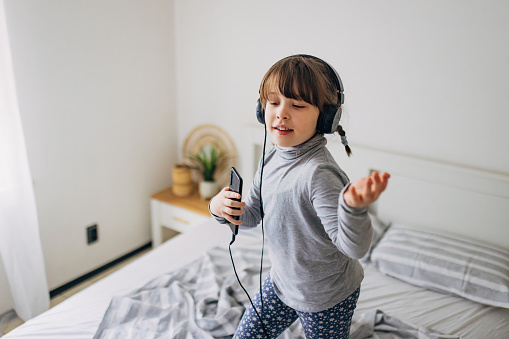 Little girl uses a mobile phone and headphones to listen to music at home. She enjoys singing, dancing and jumping on the bed during the morning hours