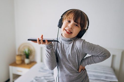 Portrait of an adorable little girl holding a mobile phone in her hands while standing on a bed, listening to music on headphones and singing