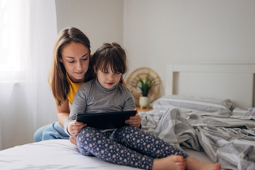 Cheerful woman takes care of her cute little daughter who is playing games on a digital tablet