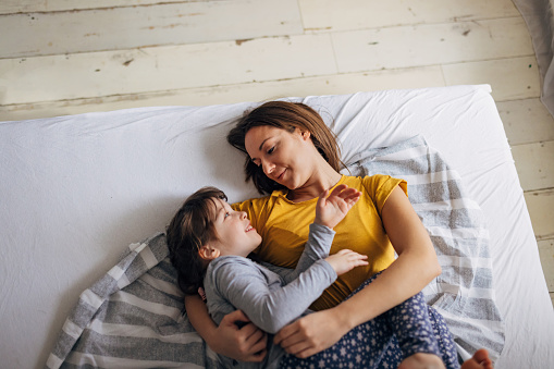 A woman and her cute little daughter are lying on a comfortable bed hugging each other and enjoying some quality mother-daughter time